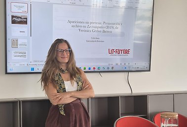 "En kvinna med glasögon står med armarna i kors framför en stor skärm som visar en PowerPoint-presentation på spanska om Verónica Gerber Biececis verk La compañía (2019). Presentationen är av Carla Alanís vid Universidad de Estocolmo inom projektet LIT-FUTURE. Rummet har röda stolar och ett bord med vattenkaraff.
