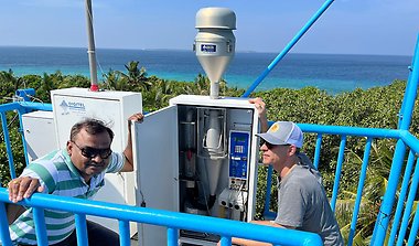 Two scientists crouch beside a measuring device, focusing on their work, with a light blue sea stretching out in the background.