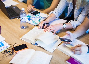 Böcker, mobiler, laptop, anteckningsblock, vattenflaska på ett bord. Studenter sitter vid bordet och man ser bara deras armar.