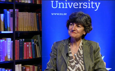 María Ángeles sitter framför en bokhylla fylld med böcker, bredvid en blå panel där texten ”University” och ”www.su.se” syns. Hon bär en grå kavaj och har en mygga fäst vid klädseln.