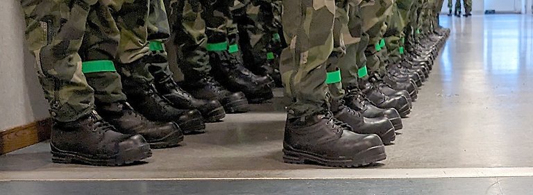 Draftees lined up in a corridor. You can see their lower legs in camouflage trousers and boots. They all have a green band around their left leg.