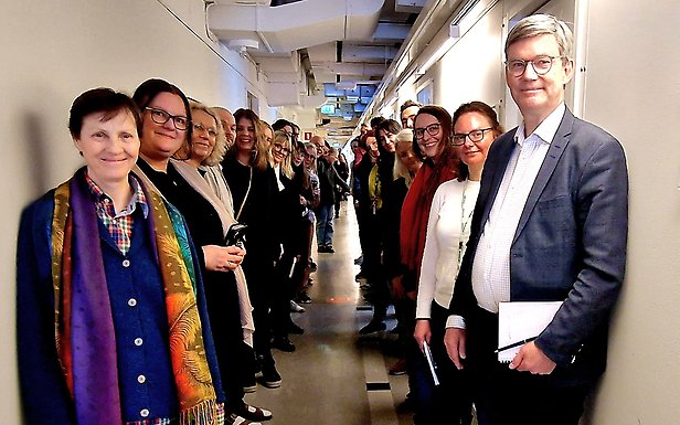 Medarbetare står i lång korridor längs väggarna och tittar mot kameran