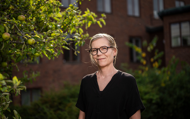 Annika Berg står bredvid ett äppelträd. I bakgrunden syns en brun tegelbyggnad i tre våningar