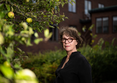 Boel Hackman står bredvid ett äppelträd. I bakgrunden syns en brun tegelbyggnad.